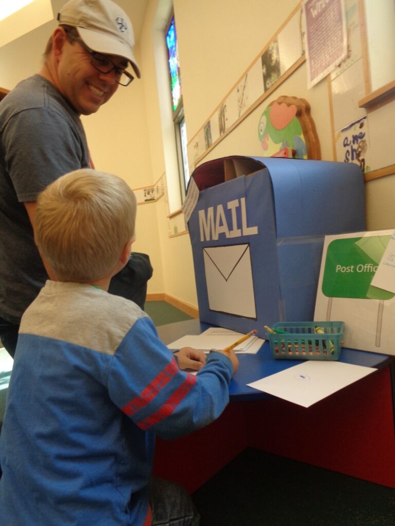 a young boy stands at a table with a cardboard box decorated like a mailbox on it; the boy is holding a pencil and looking up at his father, who is smiling back