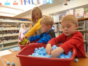two young boys in the foreground push their hands into red bins filled with bright blue fuzz balls, while an older child in the background looks at them skeptically